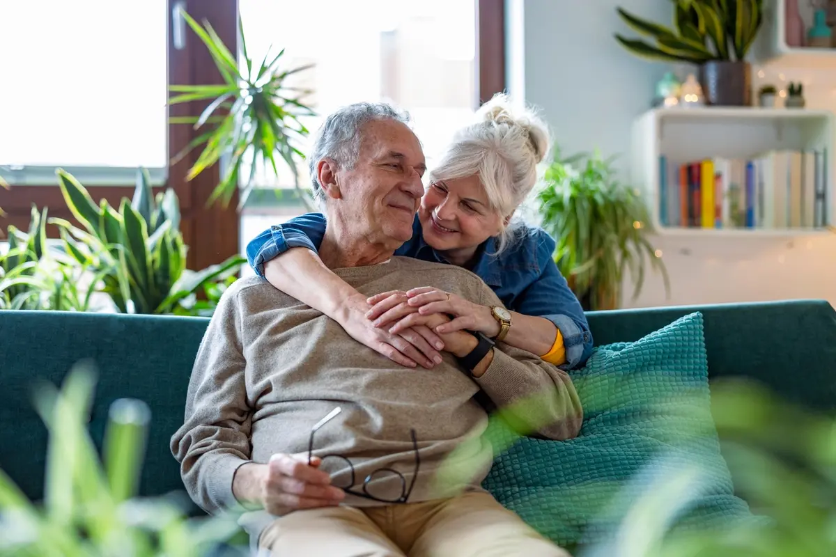 Senior couple sitting on a sofa and smiling at each other while discussing the difference between supported and assisted living