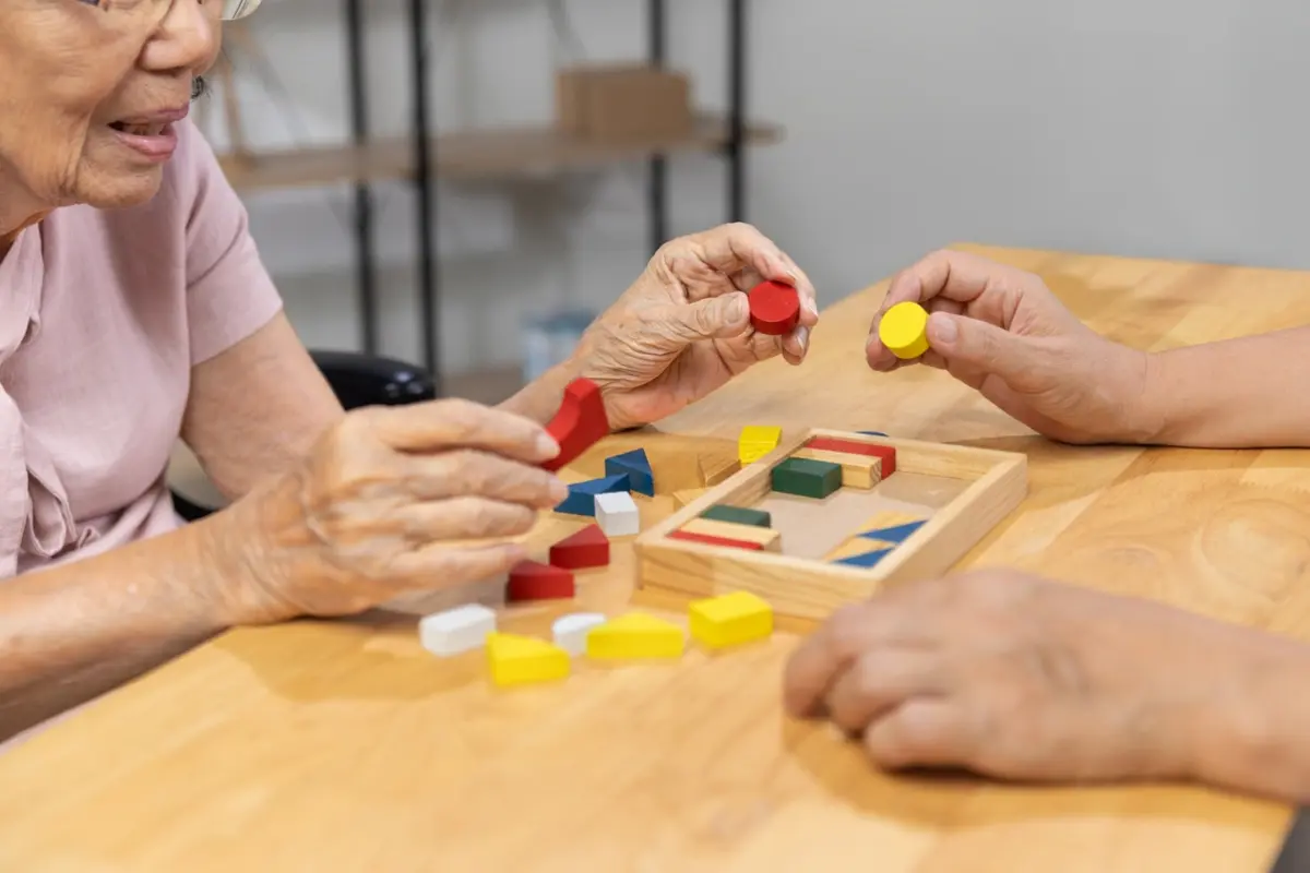 Senior woman and caregiver playing a wooden shape puzzle game to support memory and cognitive function.